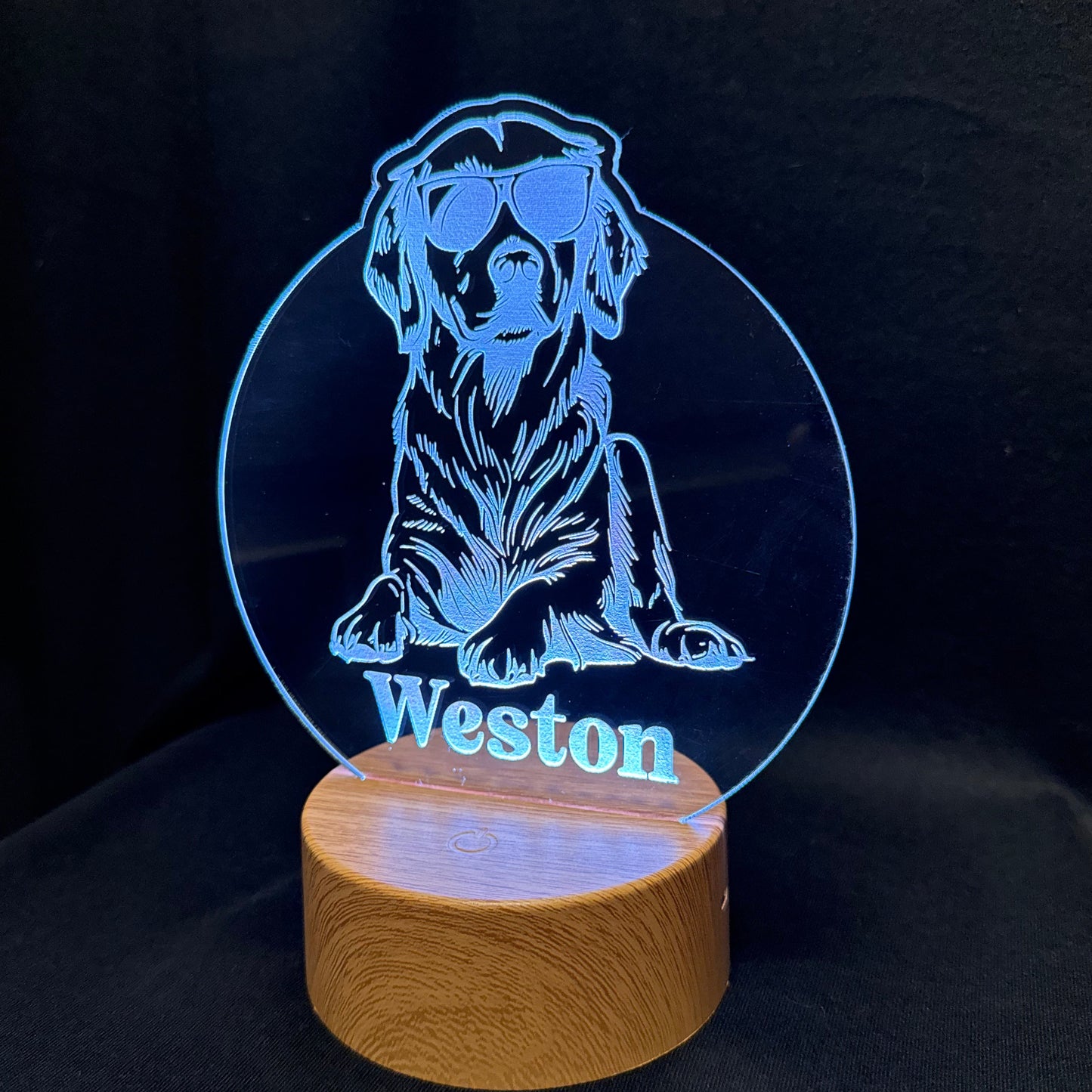 A personalized LED acrylic lamp featuring a blue laser-engraved illustration of a dog wearing sunglasses with the name 'Weston' printed below, mounted on a round wooden base against a black background.