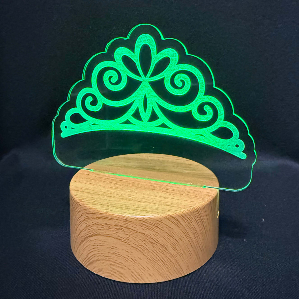 A 3D optical illusion LED lamp featuring a glowing green tiara design projected above a round, light wood-grain cylindrical base, set against a dark black fabric background.