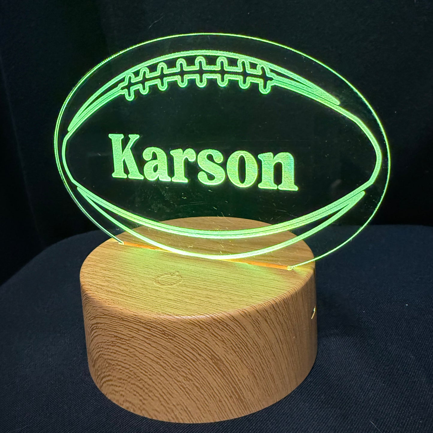 A personalized LED lamp featuring a glowing green football-shaped silhouette with the name 'Karson' printed in the center, mounted on a round natural wood base against a dark background.