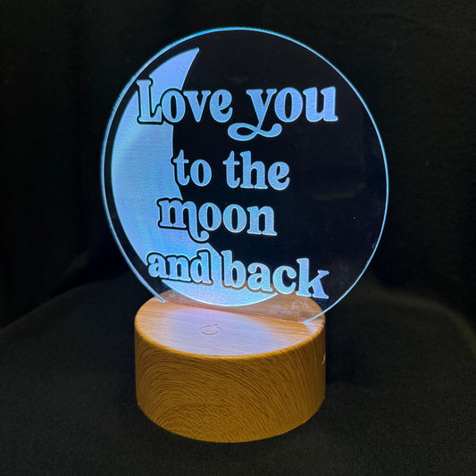 A circular acrylic LED night light with the phrase 'Love you to the moon and back' in light blue lettering, mounted on a cylindrical wooden base and photographed against a dark background.
