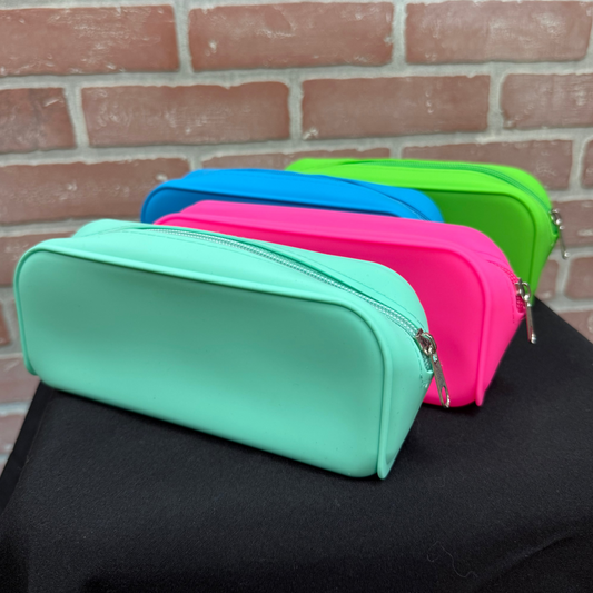 Four zippered pencil cases in mint green, pink, blue, and lime green, arranged in a row on a black cloth surface against a red brick wall background.
