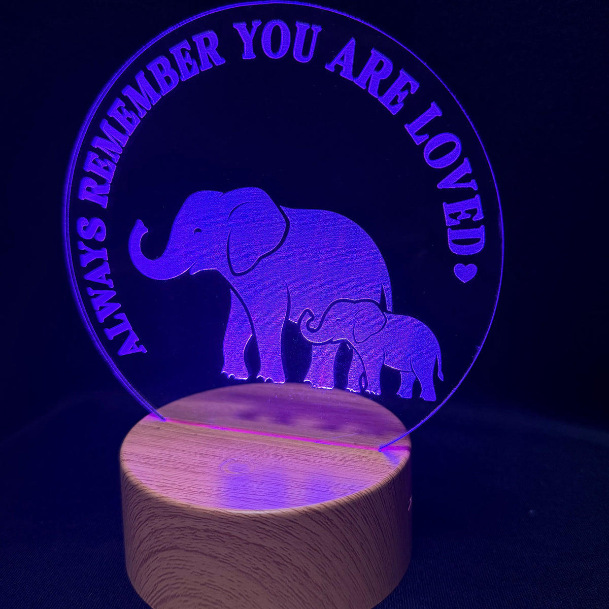 A wooden base lamp glowing with purple light, featuring an acrylic circle with the text 'ALWAYS REMEMBER YOU ARE LOVED' and silhouettes of a mother elephant and baby elephant.