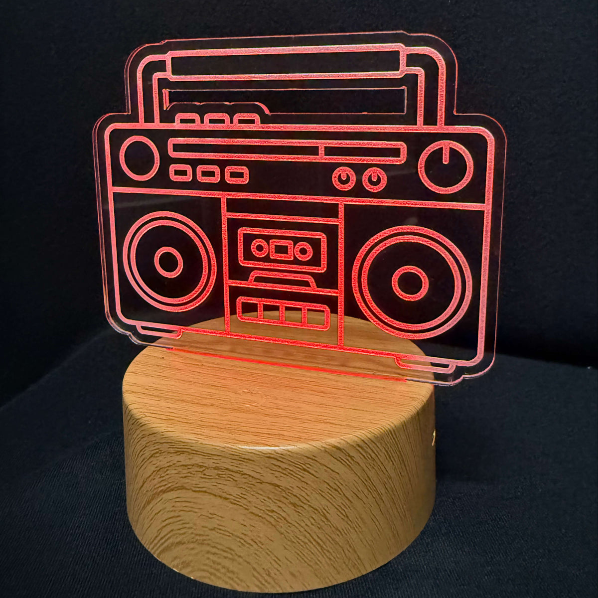 A novelty lamp featuring a red laser-engraved silhouette of a vintage boombox on a round, light-colored wooden base, set against a dark black background.