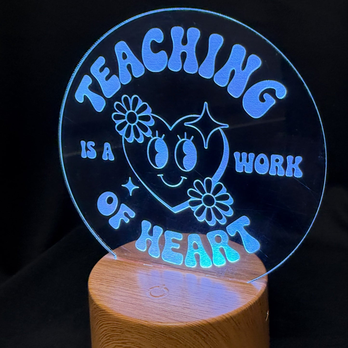 An LED acrylic 3D optical illusion lamp displaying the text 'TEACHING IS A WORK OF HEART' in glowing blue lettering on a circular clear panel, mounted on a cylindrical wooden base against a black background.