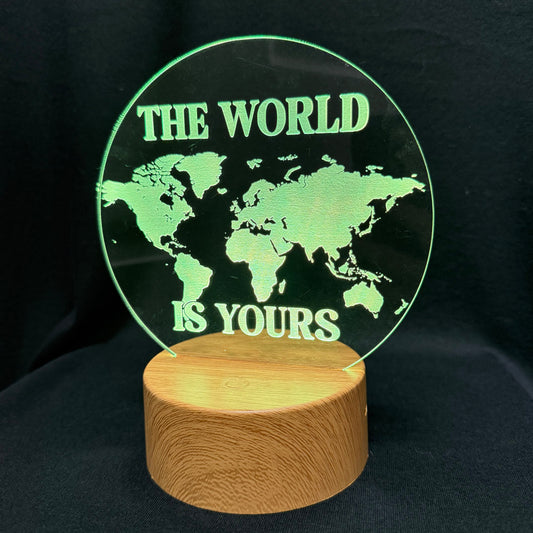 A circular clear acrylic lamp featuring a green world map design and the text 'THE WORLD IS YOURS' in green, mounted on a cylindrical light wood base against a black background.
