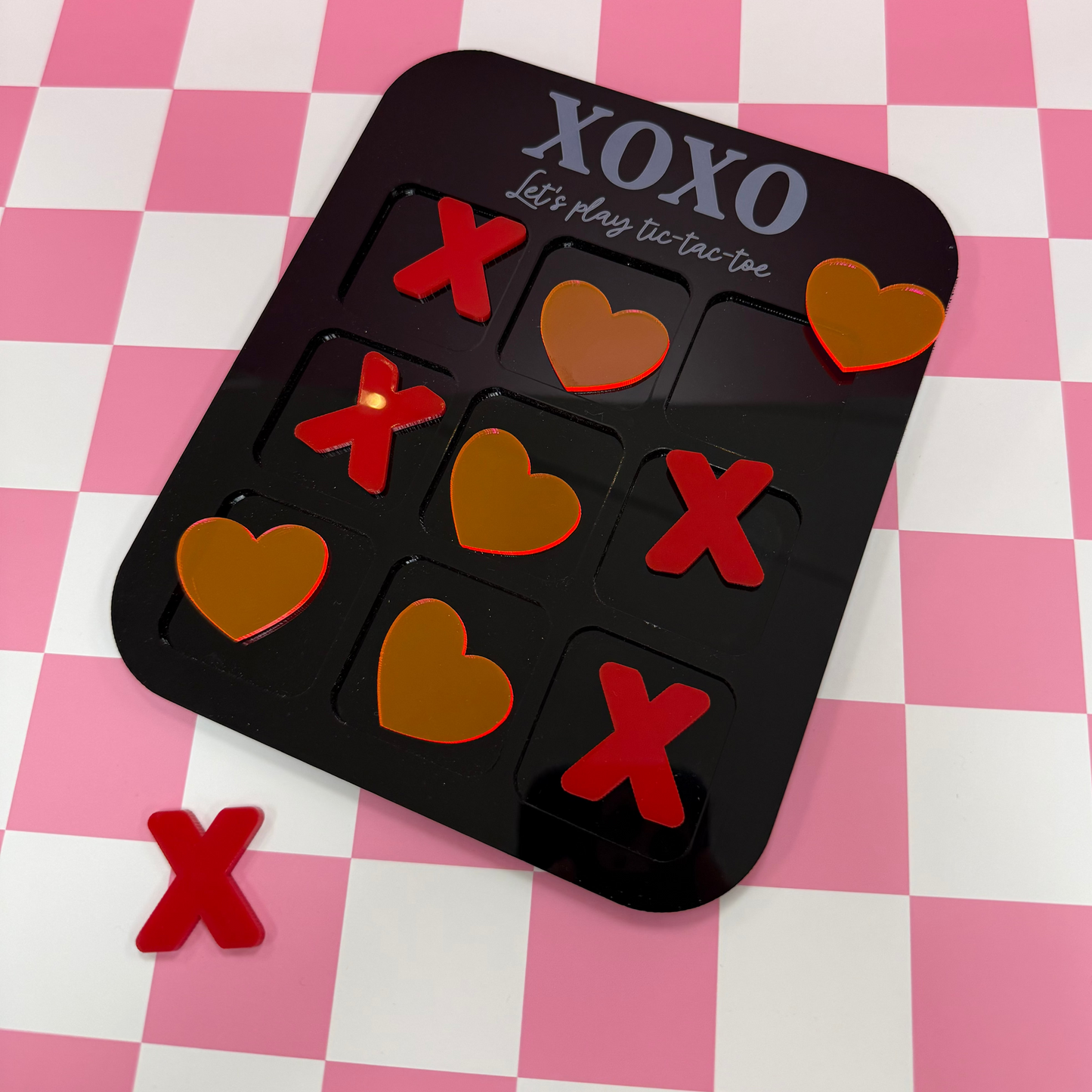 Black tic tac toe game with red game pieces that are hearts and x's on a black and white pink checkered background