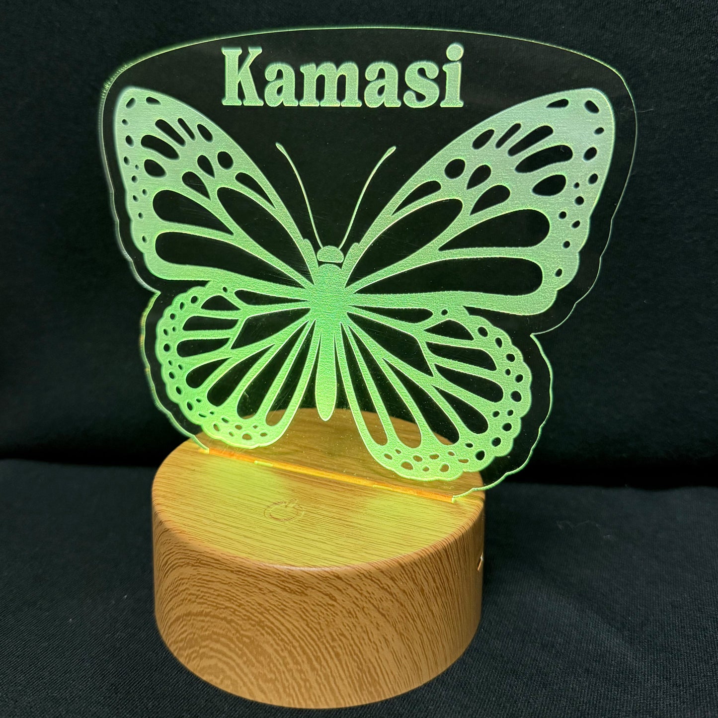 A personalized LED night light featuring a glowing green butterfly silhouette with the name 'Kamasi' engraved at the top, mounted on a round, illuminated wooden base against a black background.