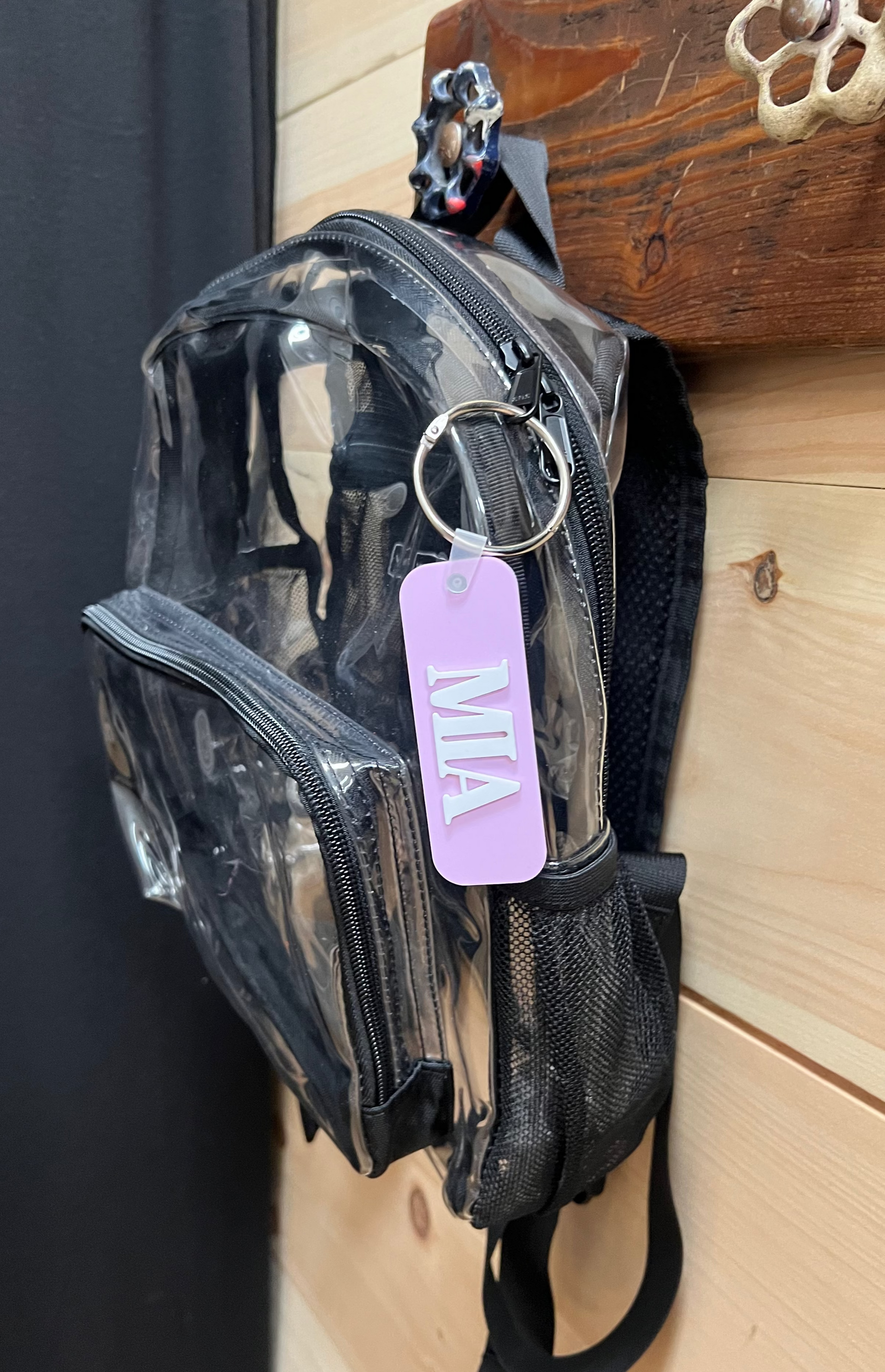 Clear transparent bag with black straps and a purple tag on a wooden surface.