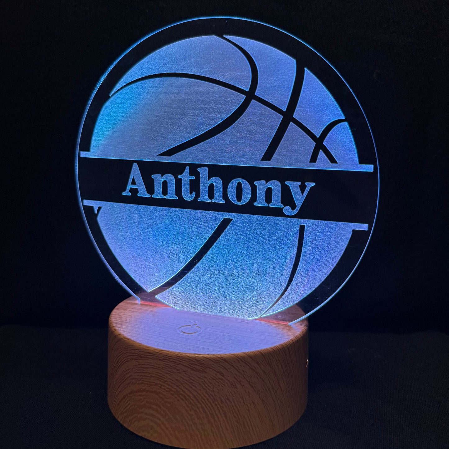A personalized 3D optical illusion lamp featuring a basketball design with the name 'Anthony' cut out, glowing with blue light on a wooden base against a black background.
