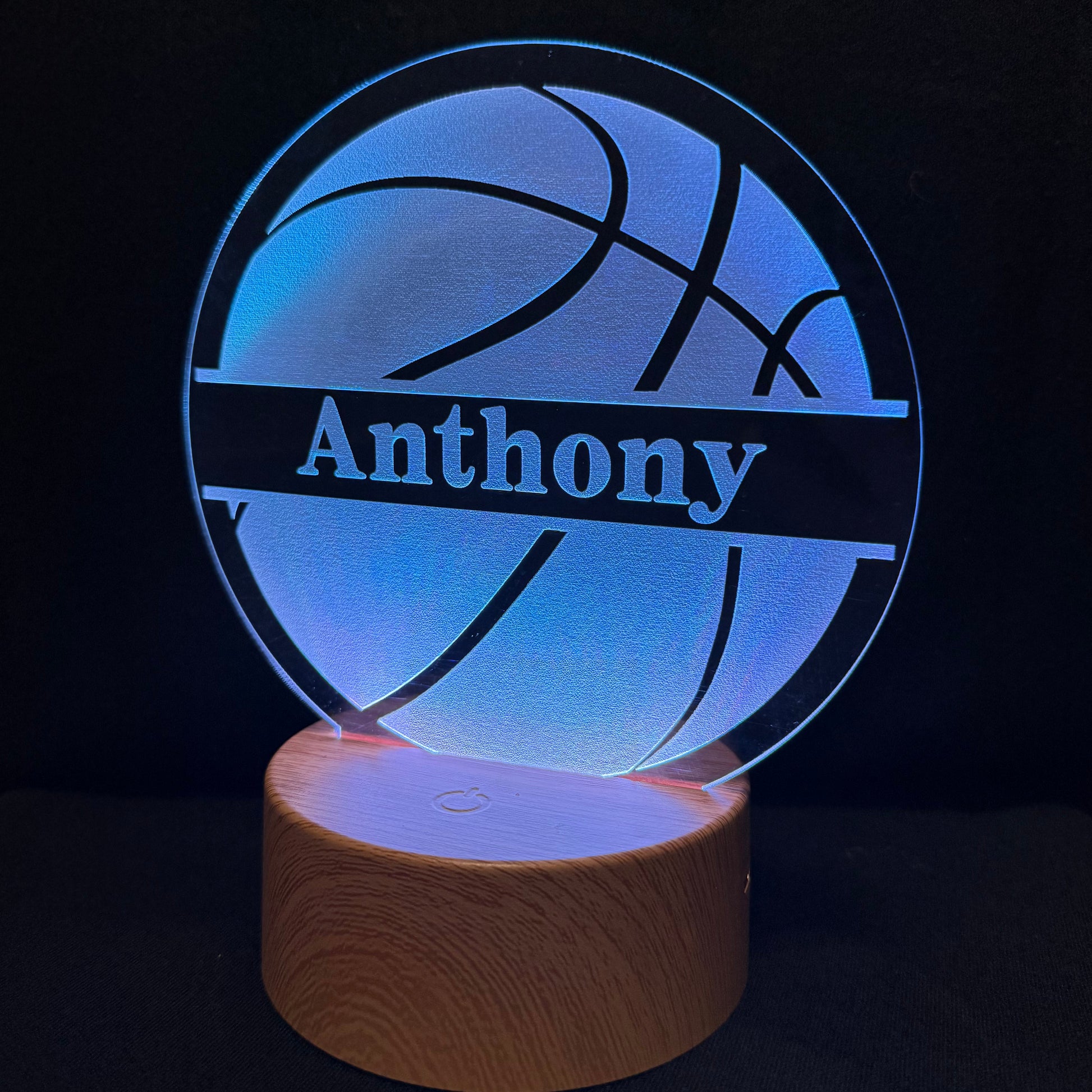 A personalized 3D optical illusion lamp featuring a basketball design with the name 'Anthony' cut out, glowing with blue light on a wooden base against a black background.