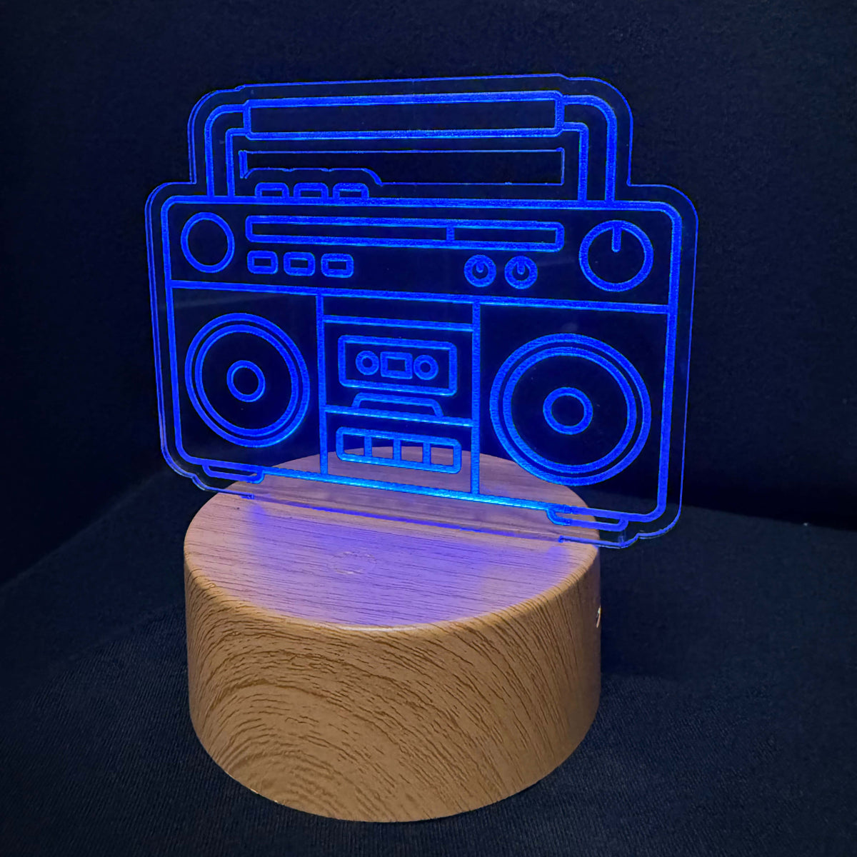 A 3D acrylic lamp shaped like a vintage boombox, glowing with a bright blue light, mounted on a cylindrical wooden base against a dark background.