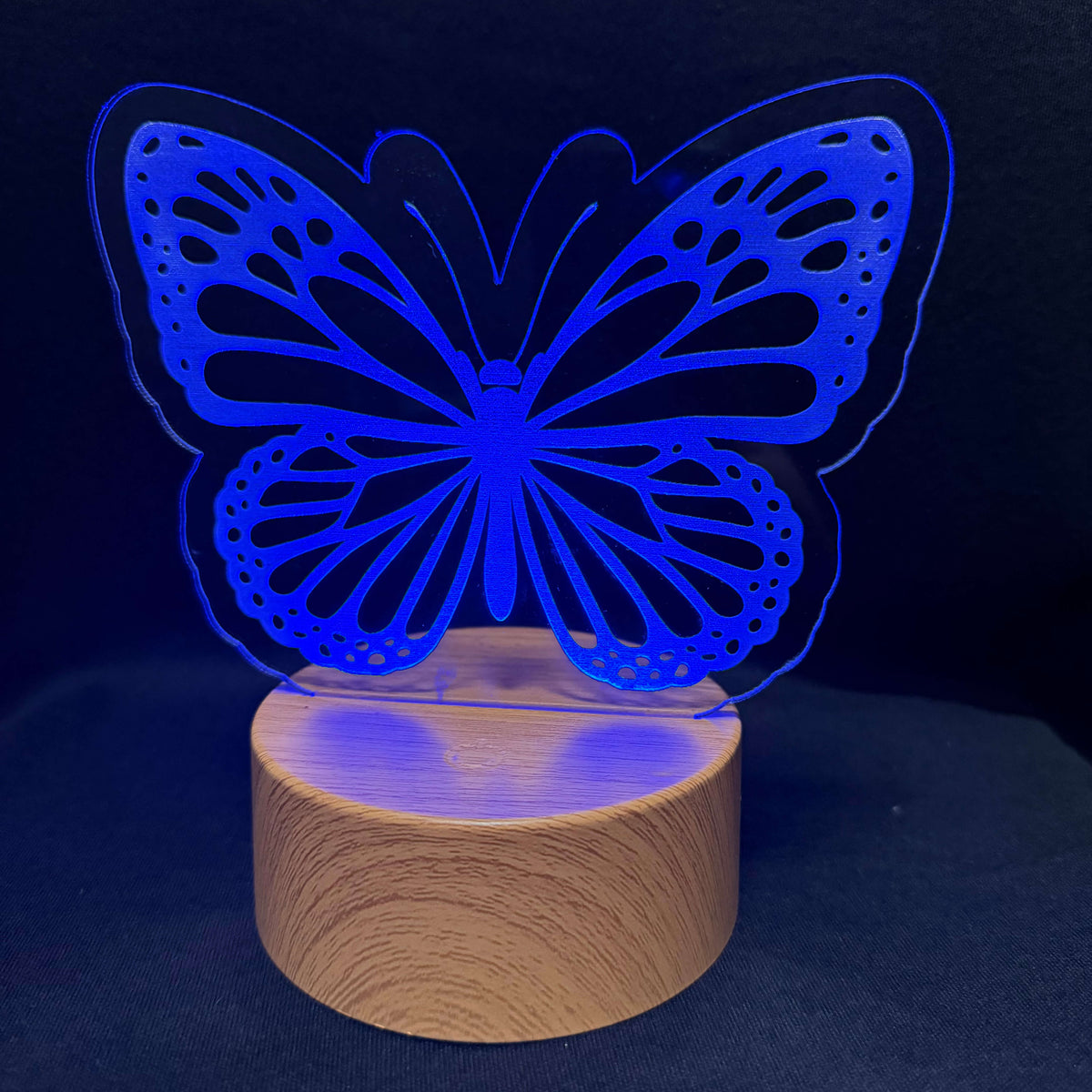 A blue LED laser-cut butterfly night light on a cylindrical wooden base, set against a dark background.