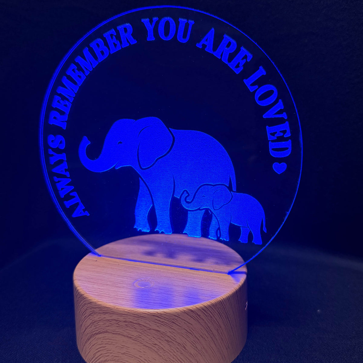 A circular acrylic LED night light with a laser-cut design of a mother elephant and her baby, surrounded by the text 'ALWAYS REMEMBER YOU ARE LOVED', mounted on a cylindrical wooden base and glowing blue against a dark background.