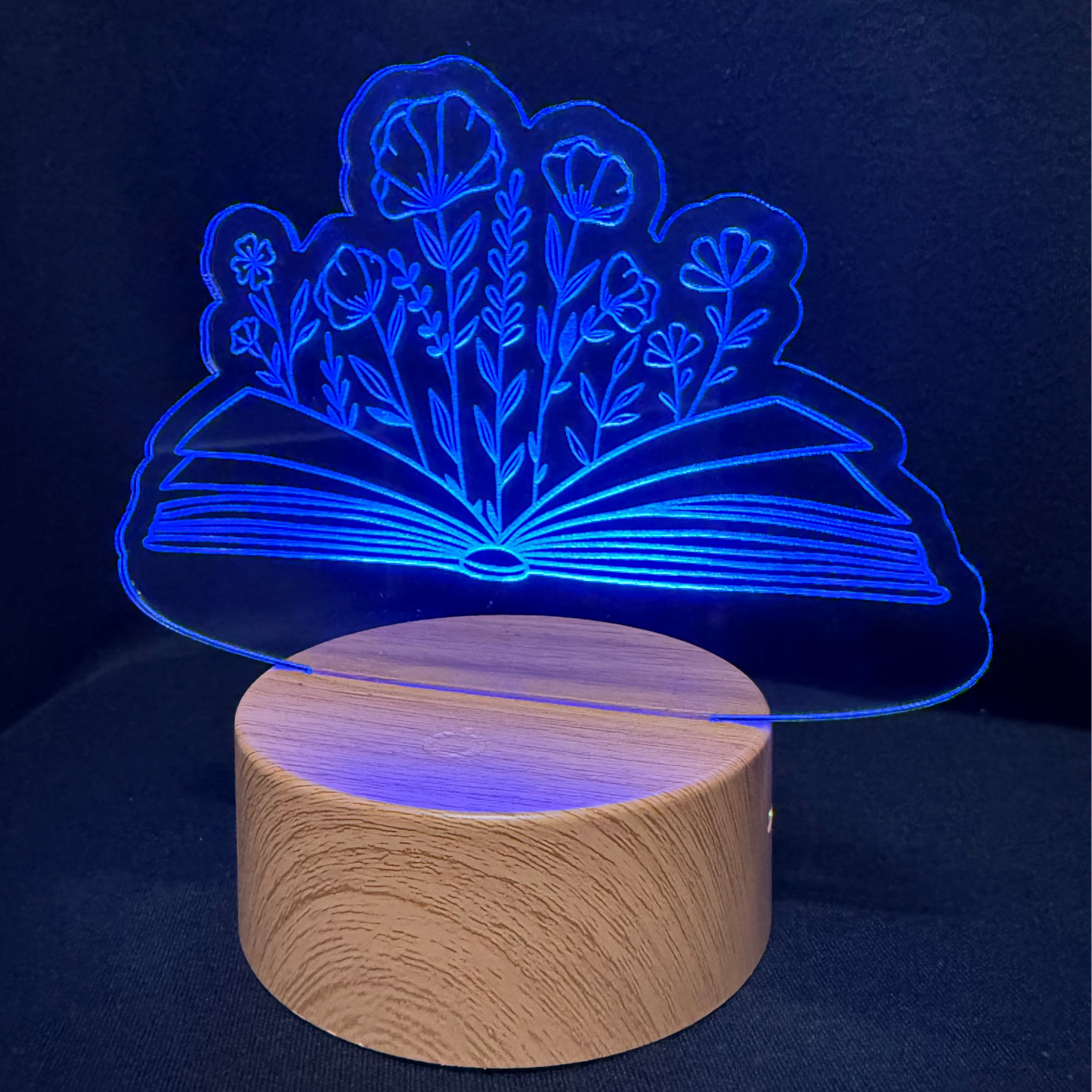 A 3D optical illusion night light shaped like an open book with glowing blue flowers, resting on a round wooden base against a dark background.