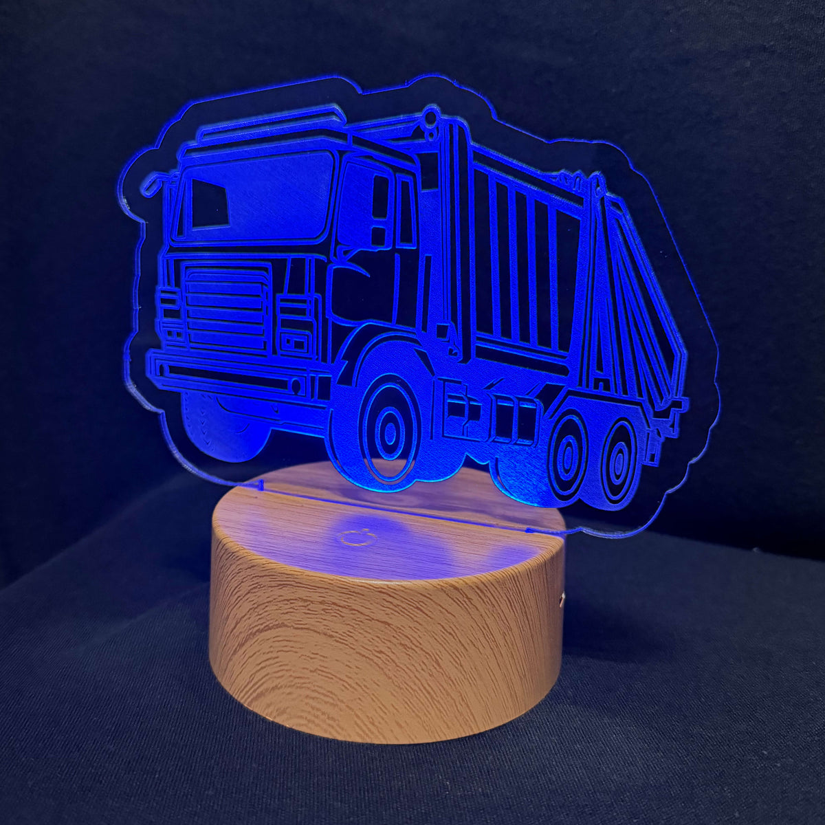 A truck-shaped LED night light with a transparent acrylic cutout of a garbage truck on a cylindrical wooden base, emitting a blue glow against a dark background.