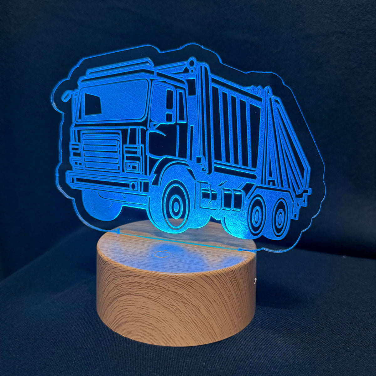 An LED acrylic night light featuring a glowing blue garbage truck silhouette on a cylindrical wooden base, set against a dark navy background.