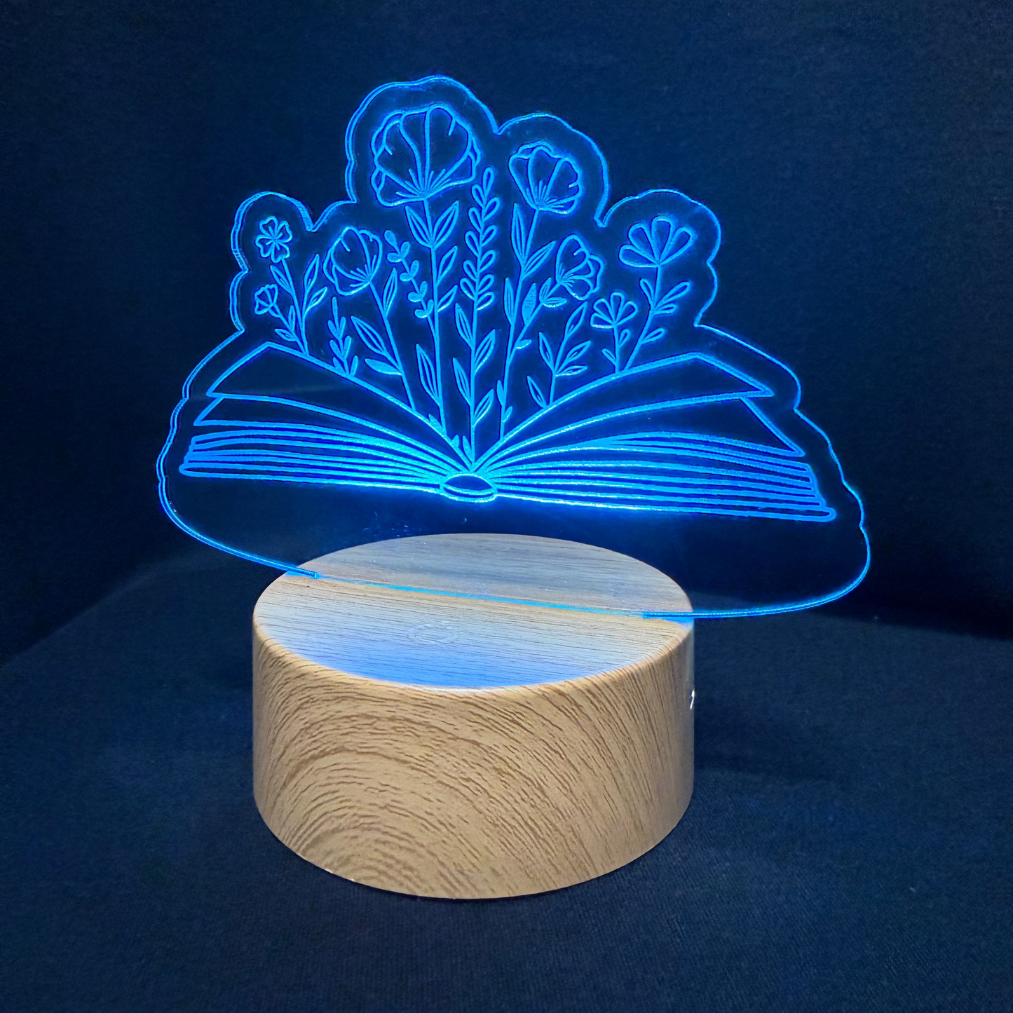 A 3D acrylic illusion night light featuring an open book with glowing blue flowers, displayed on a round light wood base against a dark navy background.