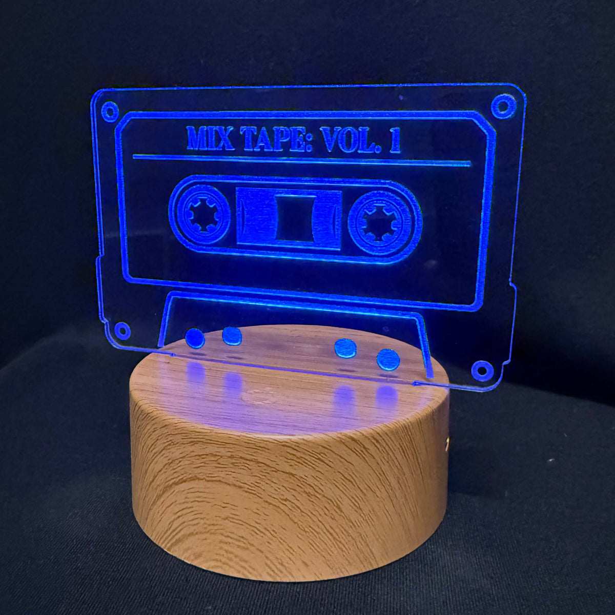 A wooden cylindrical lamp base with a transparent acrylic cassette-shaped light above it, glowing blue against a dark background. The acrylic reads 'MIX TAPE: VOL.