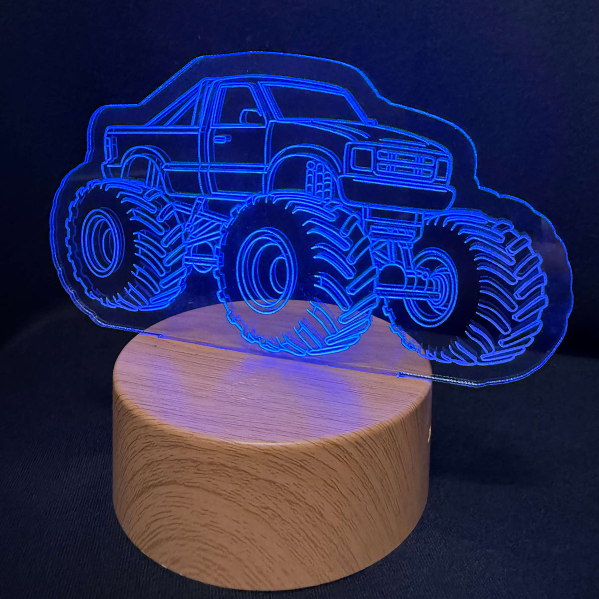 A 3D optical illusion night light featuring a glowing blue monster truck design on a round wooden base, set against a dark background.