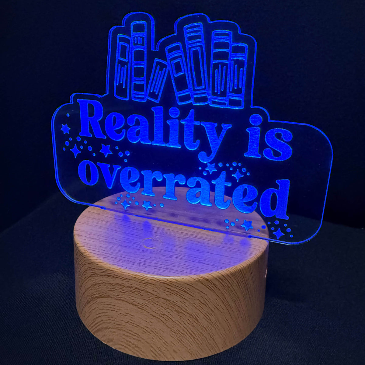An LED acrylic night light with the text 'Reality is overrated' and a bookshelf graphic, glowing blue on a round wooden base against a dark background.