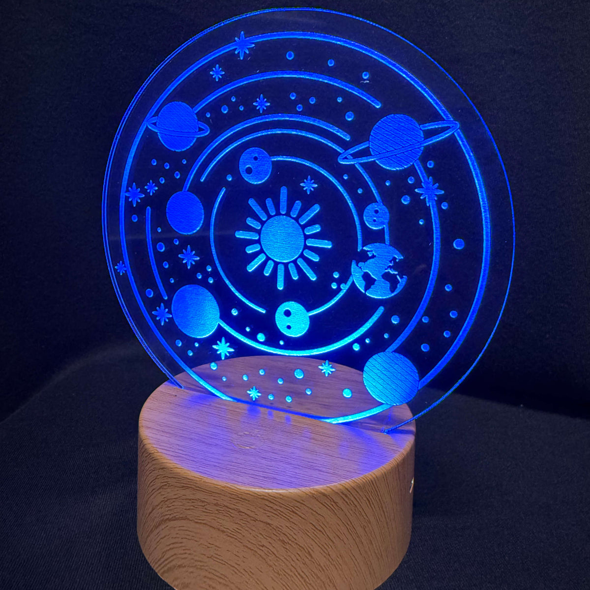 A 3D optical illusion night light featuring a glowing blue solar system design on a circular acrylic plate, resting on a cylindrical wooden base against a dark background.