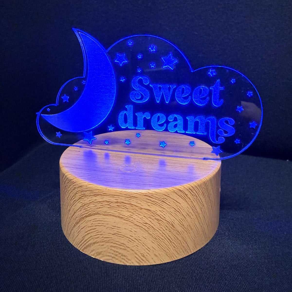 A wooden cylindrical lamp base with a glowing blue acrylic nightstand sign that reads 'Sweet dreams' with a crescent moon and stars, set against a dark background.