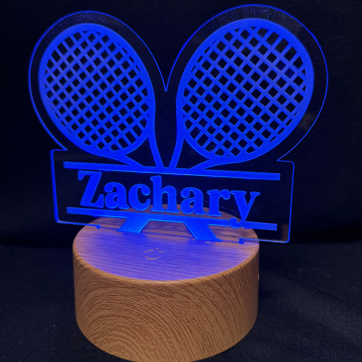 A personalized LED acrylic lamp featuring two tennis rackets and the name 'Zachary' illuminated in blue, mounted on a cylindrical wooden base against a dark background.
