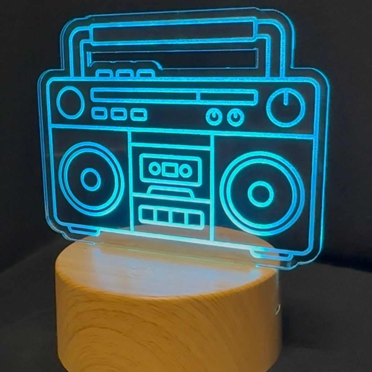 A retro boombox-shaped LED night light glowing in bright cyan blue, mounted on a cylindrical light wood base against a dark background.