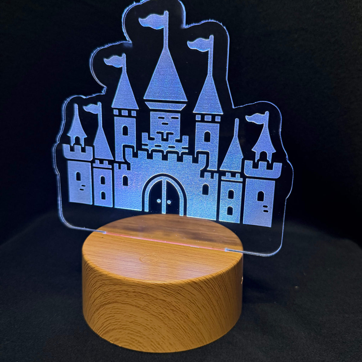A wooden cylindrical night light base supporting a clear acrylic castle-shaped panel that glows blue against a black background.