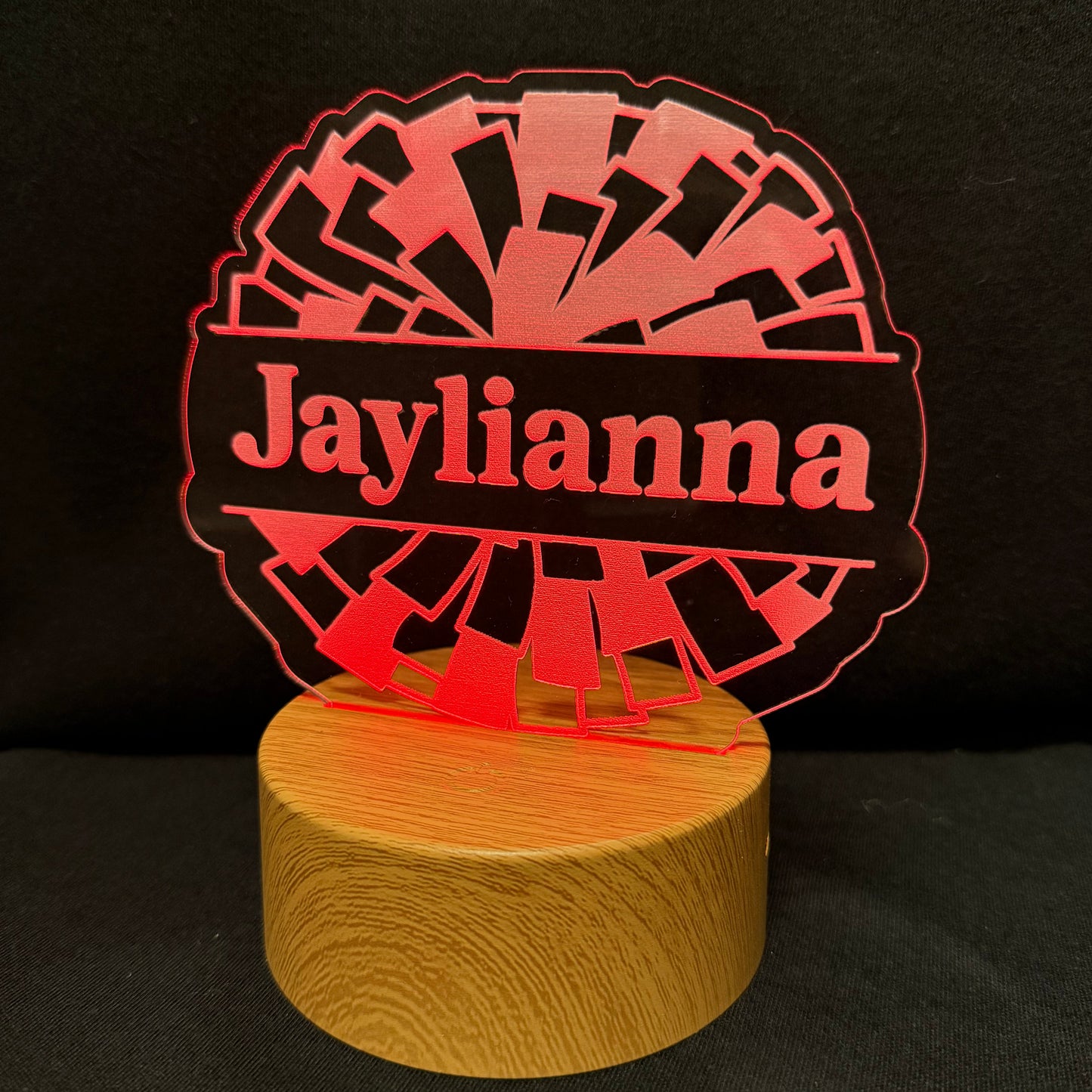 A personalized LED acrylic night light with a circular design featuring the name 'Jaylianna' in red text over a black banner, with a red geometric fan pattern behind it, mounted on a round wooden base against a black background.