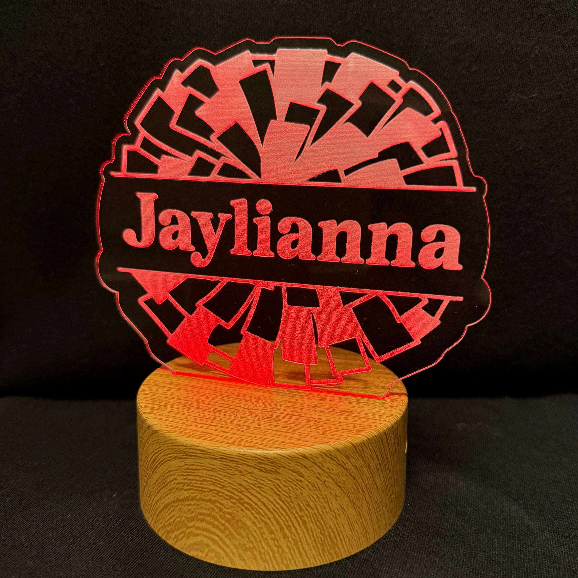 A personalized LED acrylic night light with a circular design featuring the name 'Jaylianna' in red text over a black banner, with a red geometric fan pattern behind it, mounted on a round wooden base against a black background.