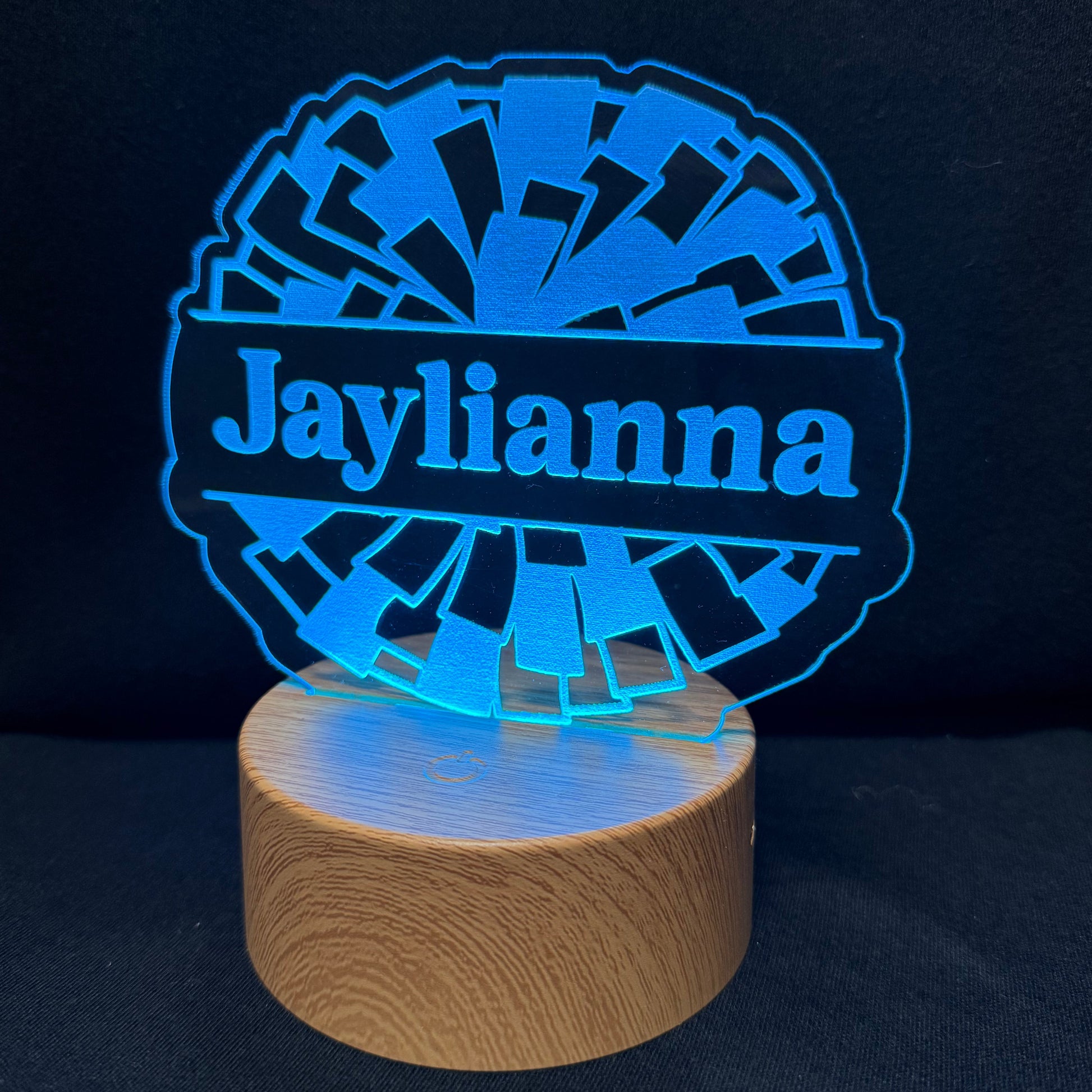 A personalized LED night light with a circular wooden base and a blue illuminated acrylic sign that reads 'Jaylianna' on a dark background.