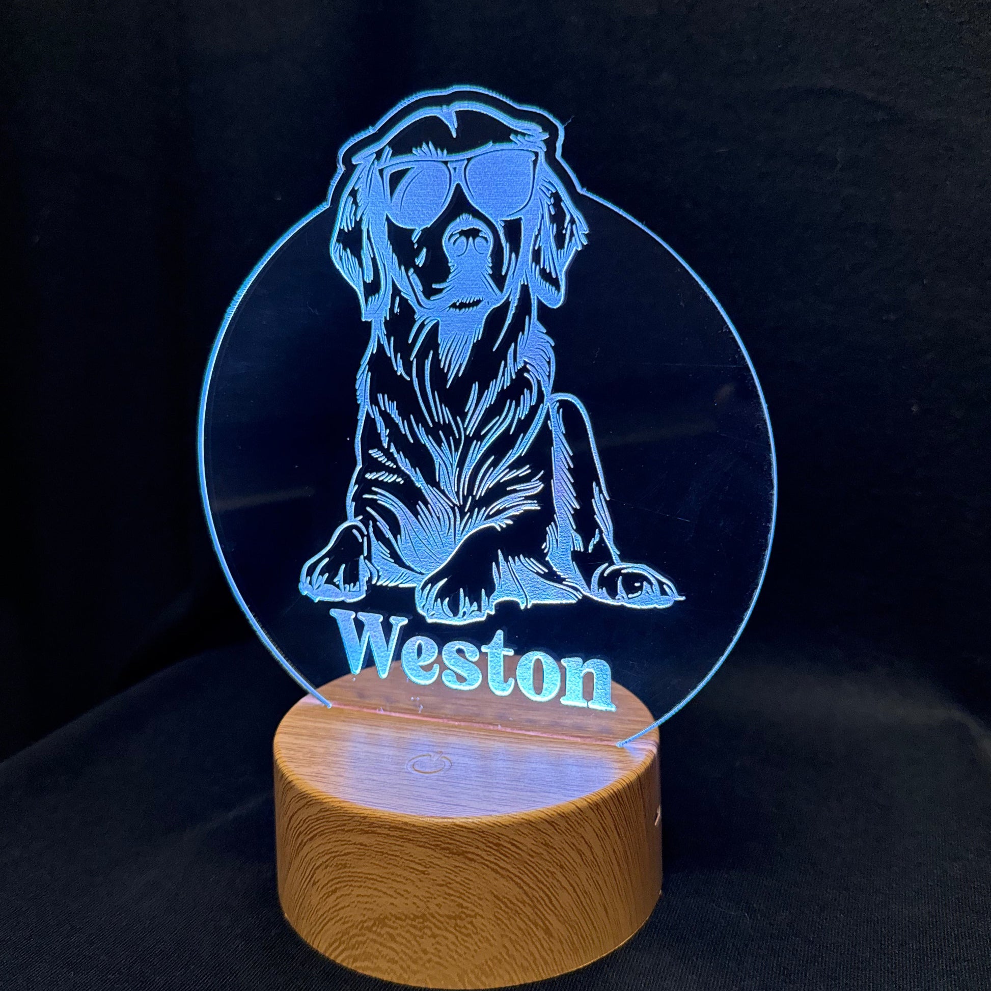 A personalized LED acrylic lamp featuring a blue laser-engraved illustration of a dog wearing sunglasses with the name 'Weston' printed below, mounted on a round wooden base against a black background.