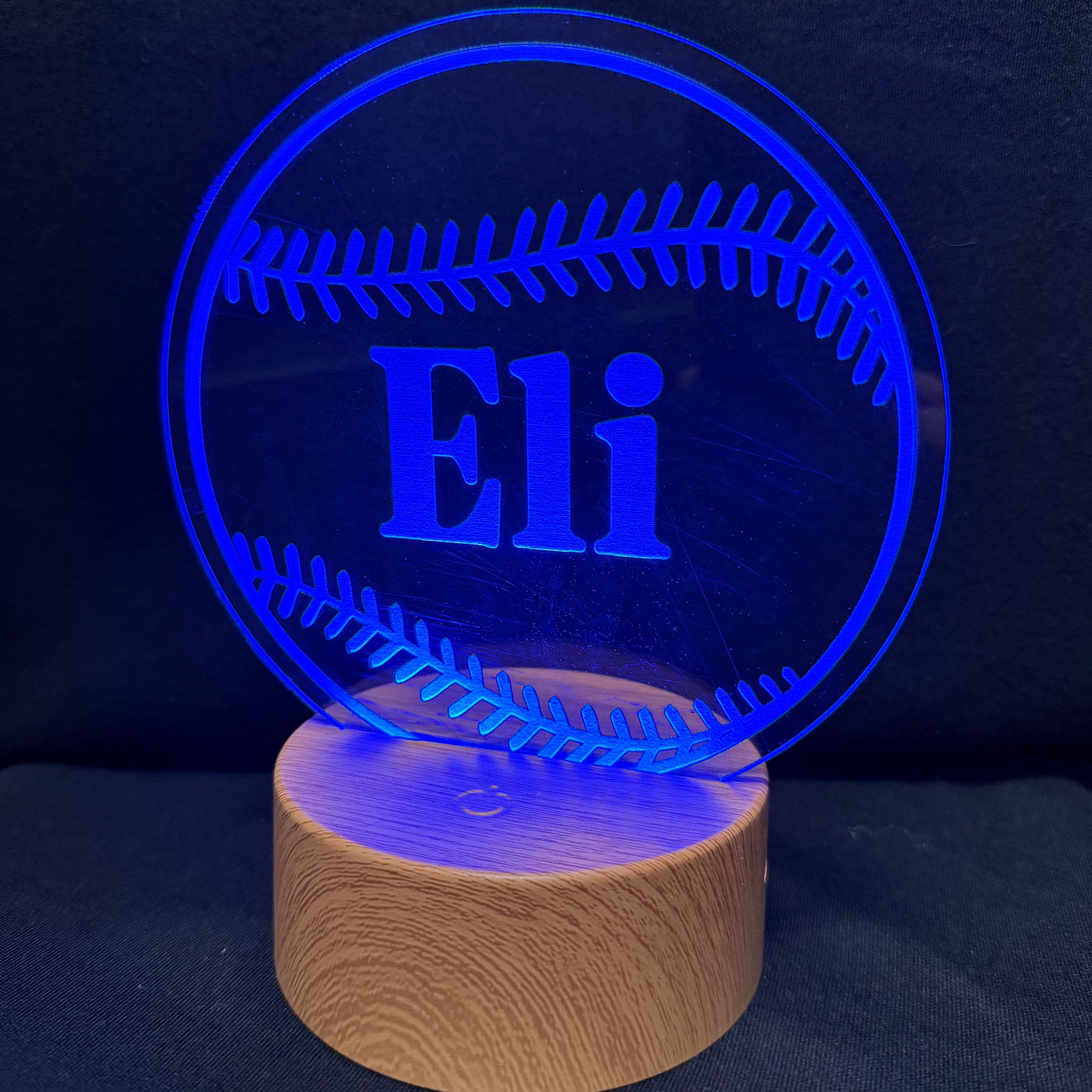 A personalized baseball-shaped LED light on a wooden base, featuring the name 'Eli' in blue illuminated acrylic against a dark background.