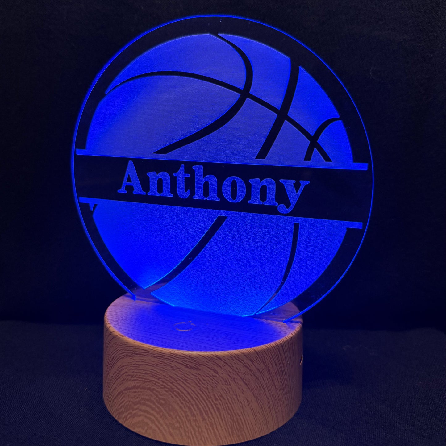 A personalized 3D optical illusion lamp featuring a basketball design with the name 'Anthony' laser-engraved on a glowing blue acrylic disc, resting on a cylindrical wooden base against a dark background.