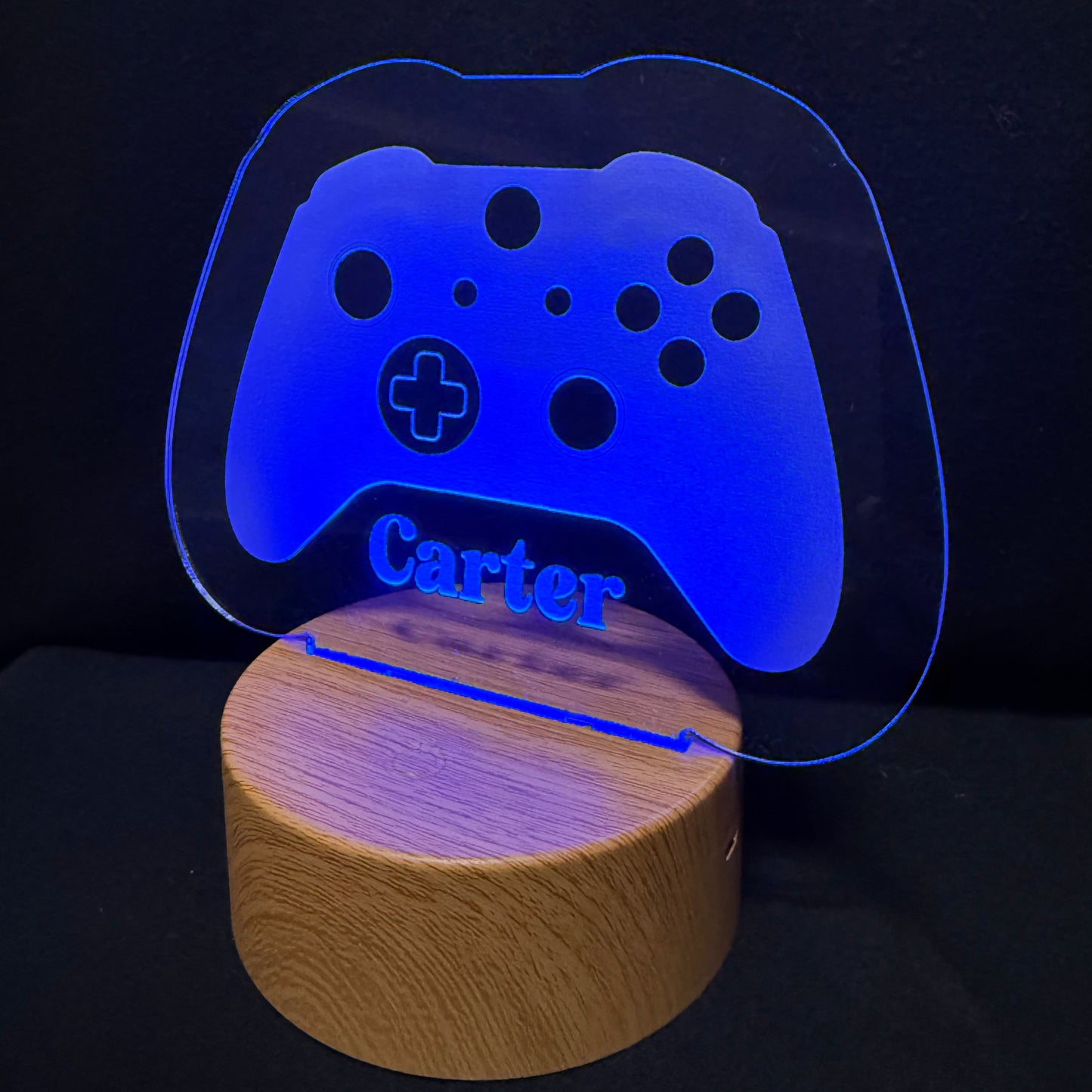 A personalized gaming-themed LED night light featuring a glowing blue translucent acrylic game controller shape with the name 'Carter' engraved, resting on a round natural wood base against a black background.