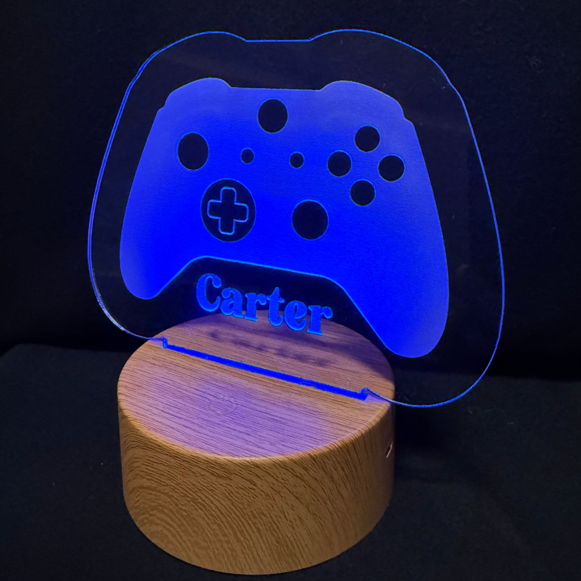 A personalized gaming-themed LED night light featuring a glowing blue translucent acrylic game controller shape with the name 'Carter' engraved, resting on a round natural wood base against a black background.