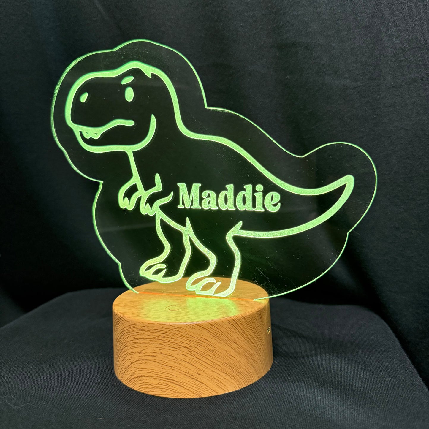 A personalized LED acrylic night light shaped like a cartoon T-Rex dinosaur, glowing green with the name 'Maddie' engraved on it, mounted on a round wooden base against a black fabric background.