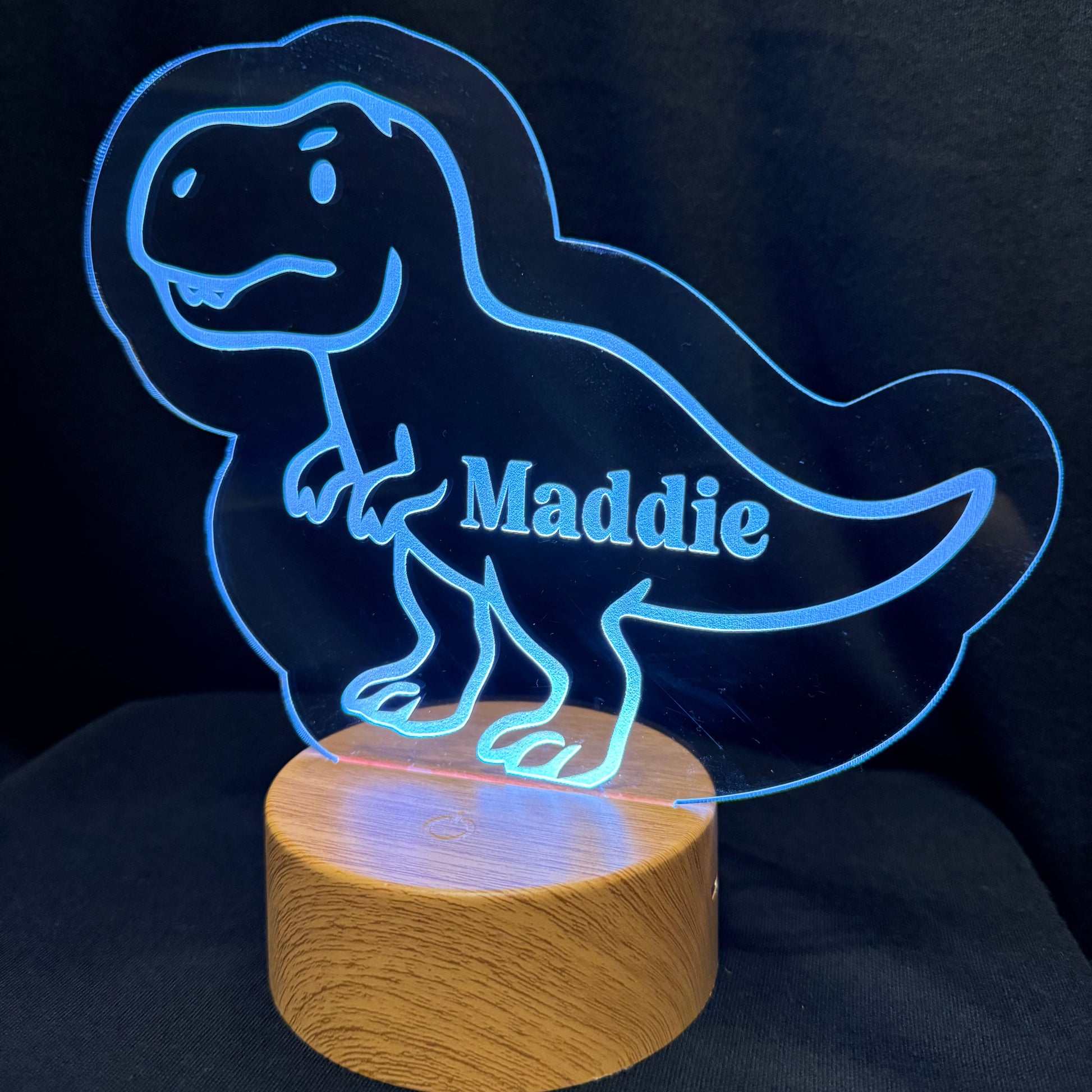 A personalized LED acrylic night light shaped like a smiling T-rex dinosaur, with the name 'Maddie' illuminated on the side, resting on a round wooden base against a dark fabric background.