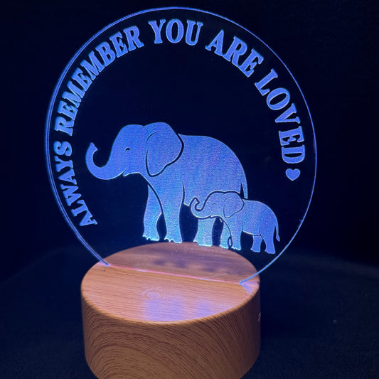 A wooden LED night light lamp featuring a glowing blue circular acrylic panel with an elephant and baby elephant silhouette and the text 'ALWAYS REMEMBER YOU ARE LOVED', set on a cylindrical wooden base against a dark background.