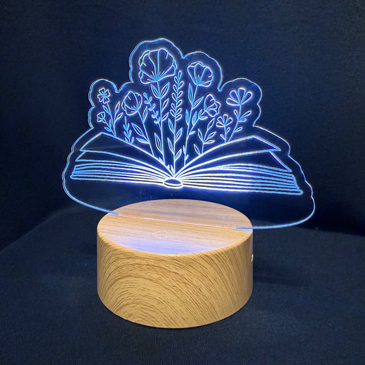 A 3D acrylic LED night light shaped like an open book with glowing blue flowers, resting on a round wooden base against a dark background.