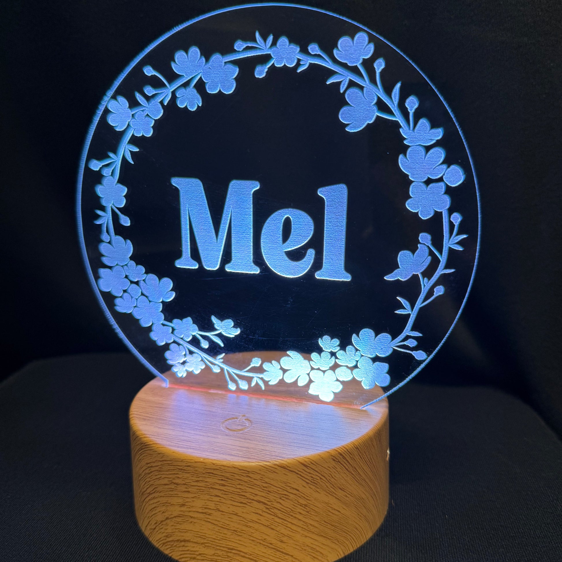 A personalized LED night light with a circular clear acrylic top featuring the name 'Mel' in blue, surrounded by a floral wreath design, mounted on a cylindrical wooden base that glows with a warm light against a black background.