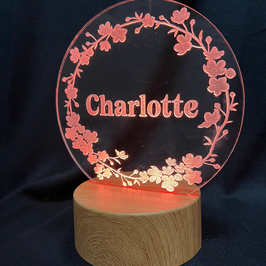 Personalized Floral Circle Night Light featuring a laser-engraved acrylic panel with a delicate floral wreath surrounding a custom name, illuminated by a warm LED light on a wooden base.