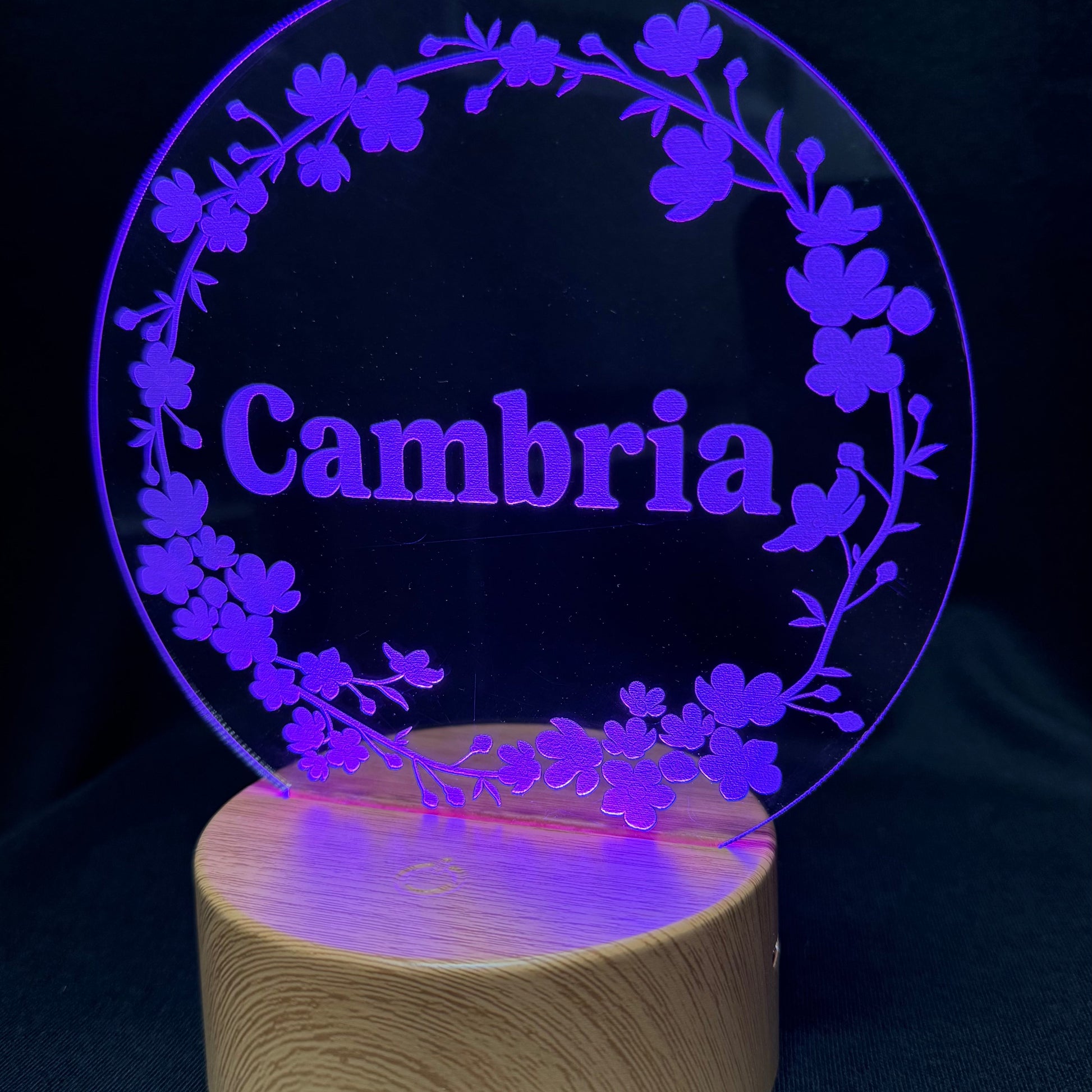 A personalized acrylic LED night light featuring a circular clear panel with a floral wreath design and the name 'Cambria' in the center, illuminated in purple light and mounted on a cylindrical wooden base against a dark background.