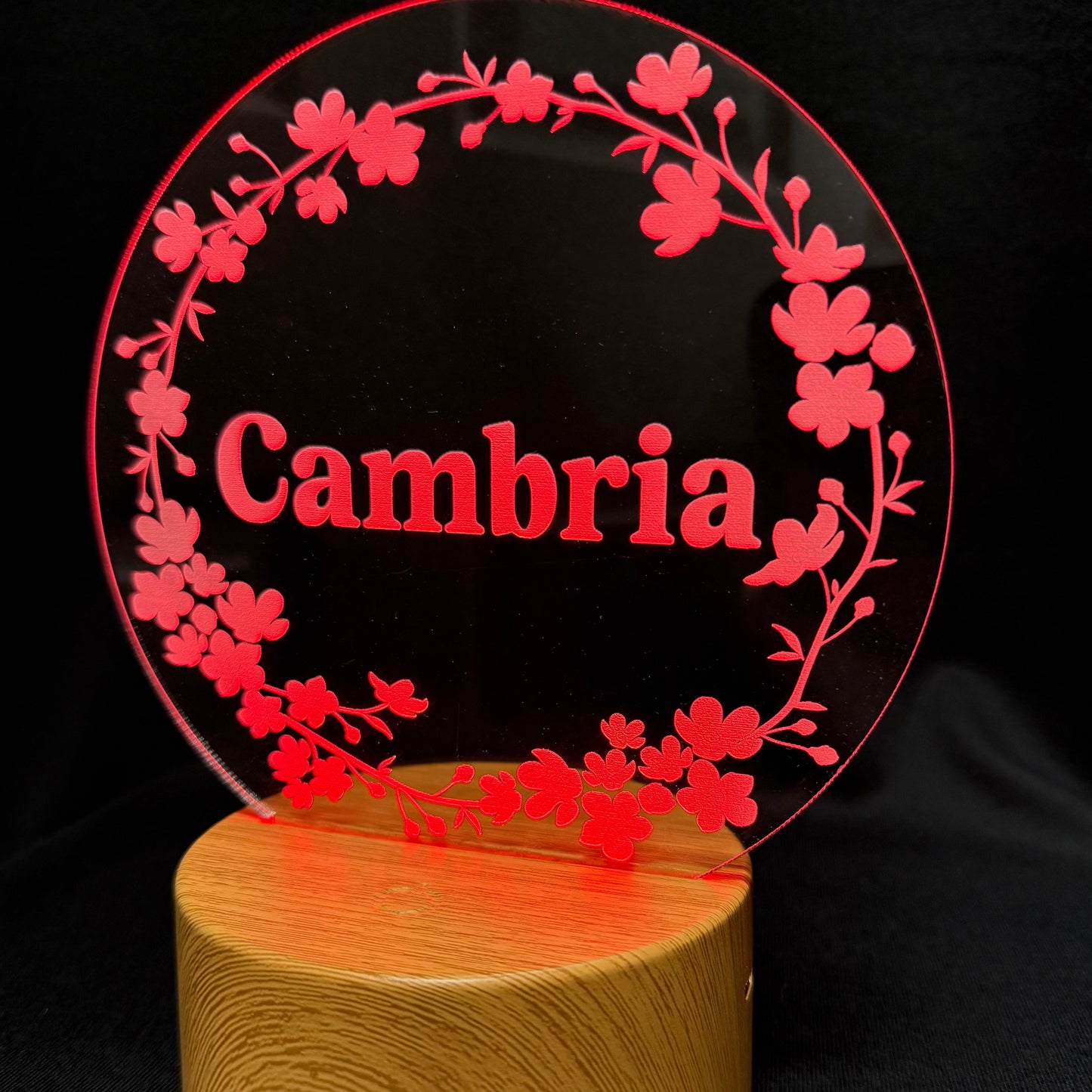 A personalized acrylic plaque with a red cherry blossom wreath and the name 'Cambria' in red text, mounted on a cylindrical wooden base against a black background.