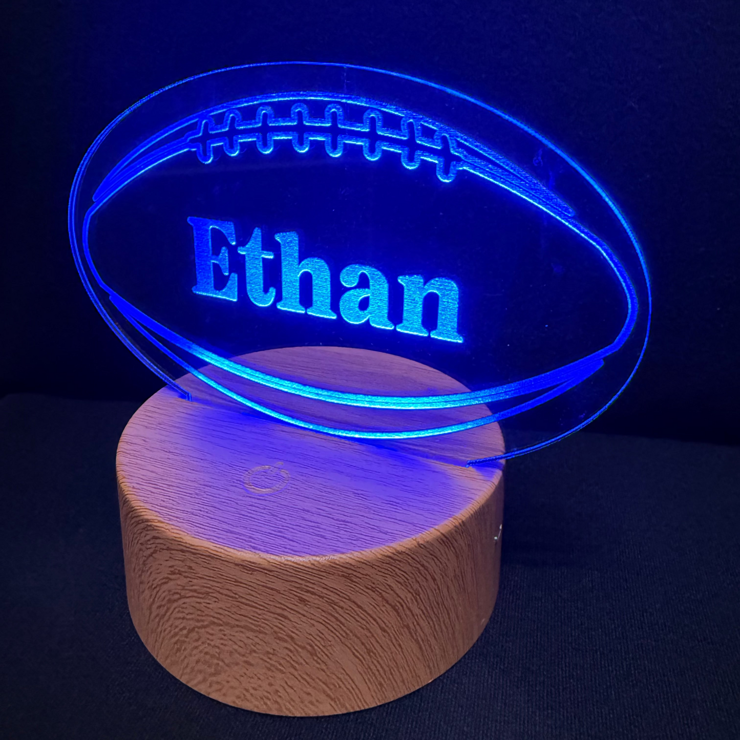 A personalized LED lamp featuring an engraved football-shaped acrylic plate with the name 'Ethan' illuminated in blue, resting on a round wooden base against a dark background.