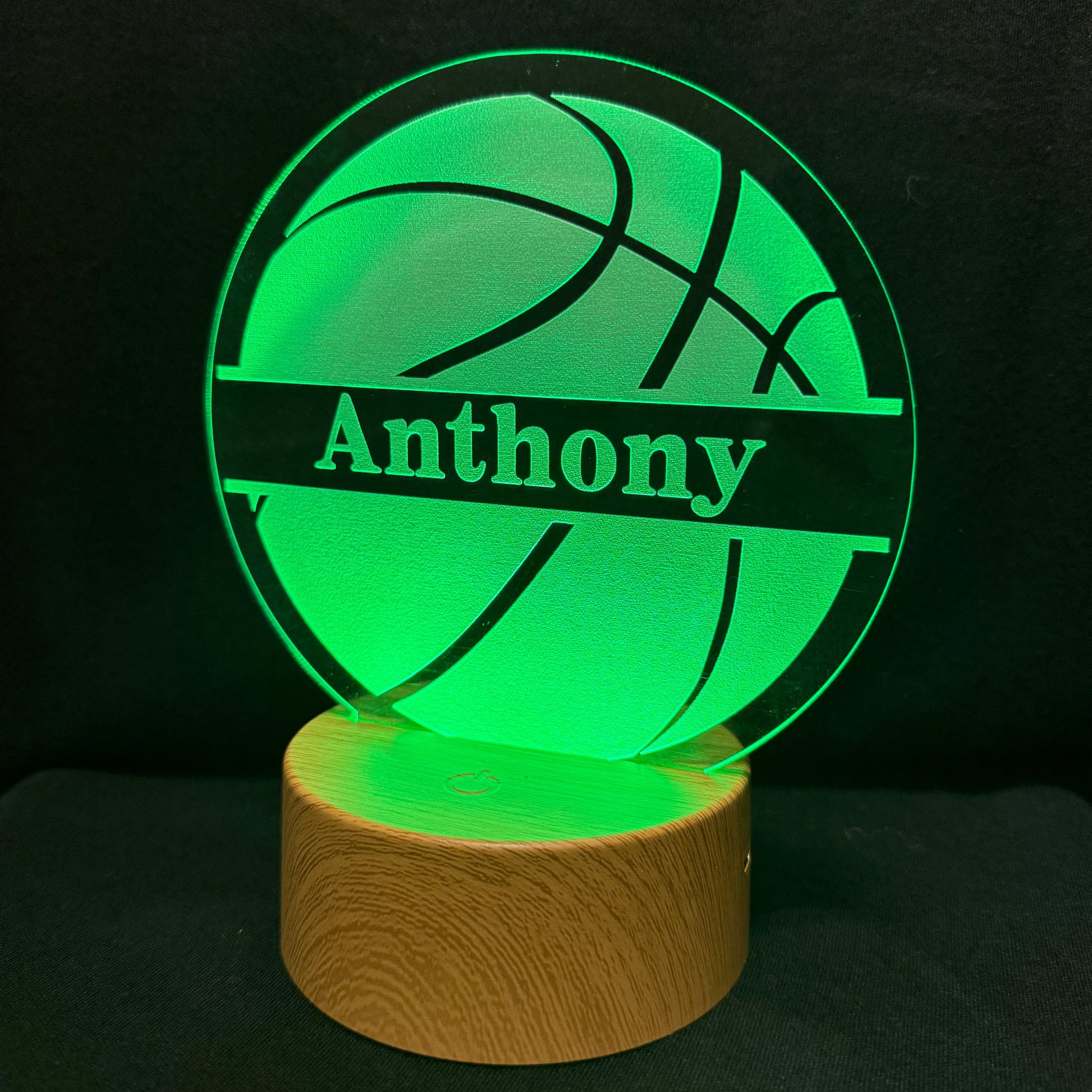 A personalized 3D acrylic LED lamp shaped like a basketball, glowing bright green with the name 'Anthony' laser-engraved across the center, resting on a cylindrical wooden base against a dark background.
