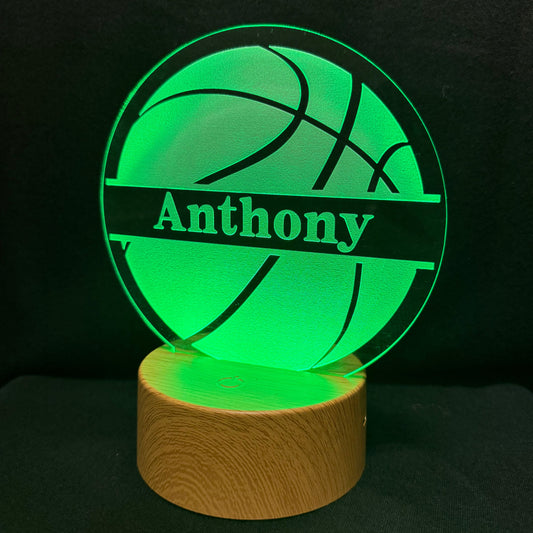 A personalized 3D acrylic LED lamp shaped like a basketball, glowing bright green with the name 'Anthony' laser-engraved across the center, resting on a cylindrical wooden base against a dark background.