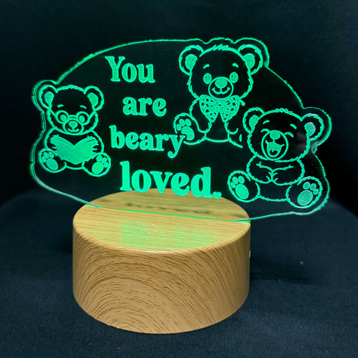 A 3D laser-engraved LED night light featuring three cute teddy bears and the text 'You are beary loved.' glowing in green, mounted on a round wooden base against a dark background.
