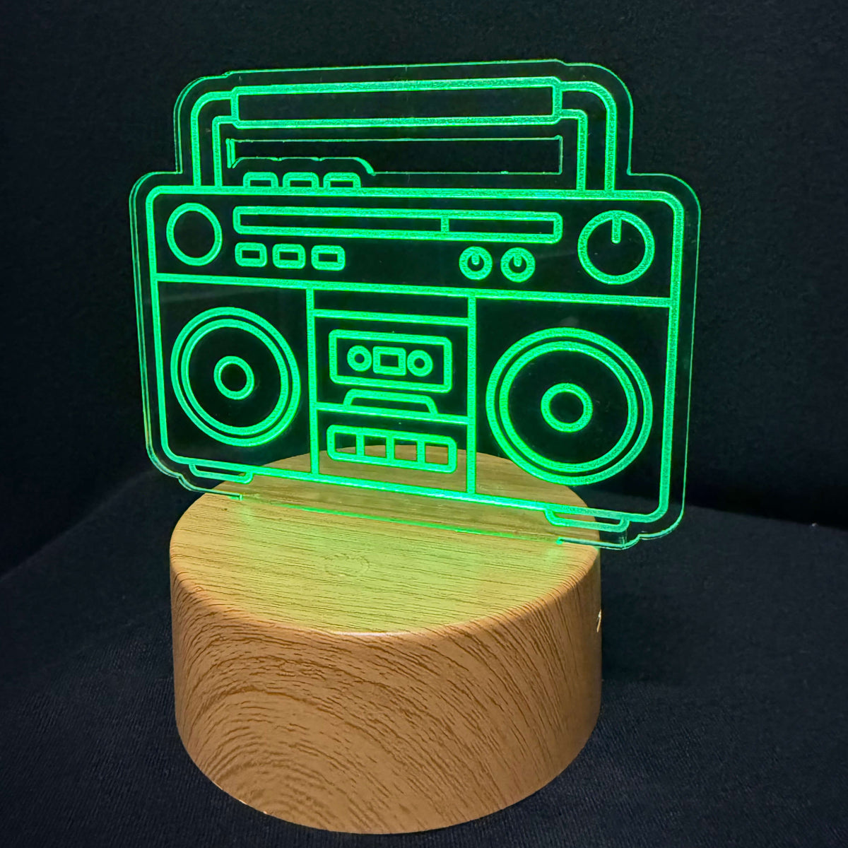 An LED acrylic lamp featuring a glowing green boombox design resting on a cylindrical light wood base, set against a dark black background.